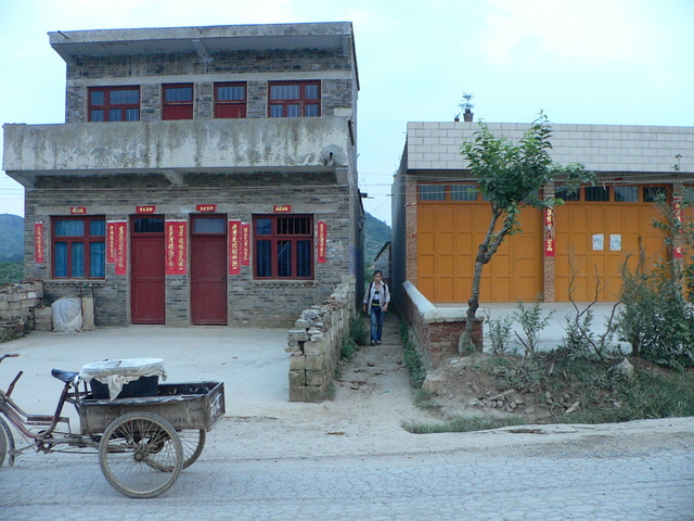 Ah Feng emerges onto the main road between two buildings, with a bean curd hawker's tricycle parked out front. The confluence is 470 metres SSE.