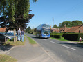 #10: A double decker bus drives along Stockton-on-the-Forest's one street.  People are gathering to watch the "Tour of Britain" bike racers.